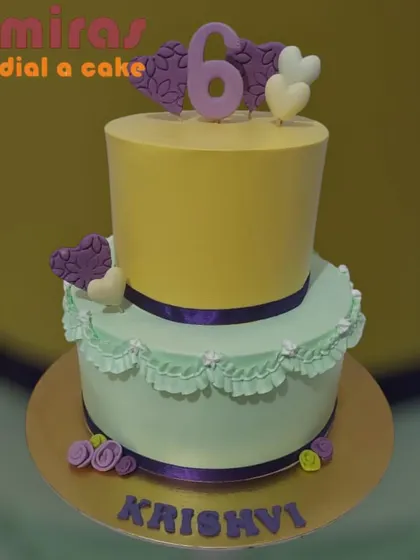 A sweet and simple two-tier cake for a 6th birthday. The pastel yellow and mint green layers are decorated with purple hearts and delicate ruffles, creating a soft and pretty look.