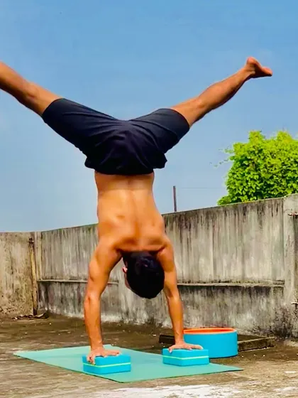 A powerful handstand with legs in a wide split, performed outdoors using yoga blocks for elevation and support.