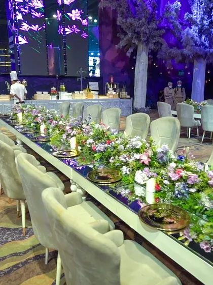 A long, beautifully decorated table set for a VIP dinner. The mirrored surface reflects the floral centerpieces and ambient lighting, creating an elegant and intimate setting for a multi-course gourmet meal.