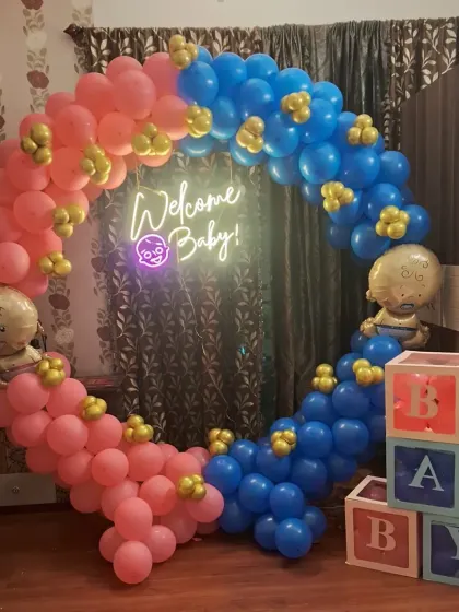 A well-lit photo of a pink and blue circular balloon arch for a gender reveal party. The setup includes baby foil balloons and 'BABY' blocks, perfect for photos.