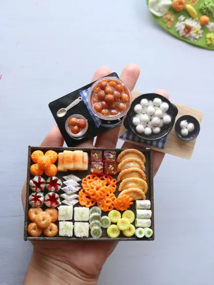 A hand holding a collection of sweet miniatures, including gulab jamun, rasgulla, and a box of assorted sweets.