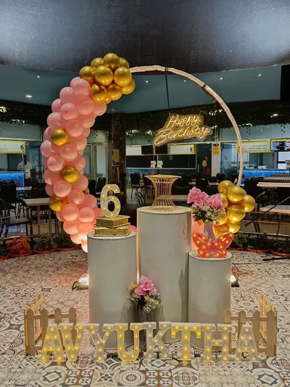 A sophisticated birthday setup in a restaurant or hall. The decor features a semi-circular balloon arch in pink and gold, multiple pedestals for cake and desserts, and marquee letters.