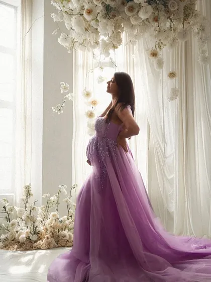 A dreamy, light-filled portrait. The mom-to-be stands by a window in a lavender gown, surrounded by a cascade of white flowers.