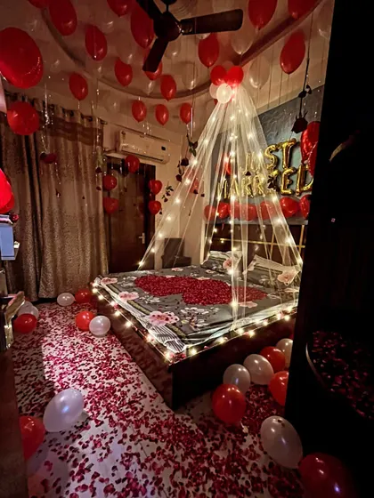 Here is a full view of a romantic room setup, showing the floor completely covered in red rose petals. This design includes a fairy light canopy over the bed, 'Just Married' balloons, and dozens of red and white balloons on the ceiling.