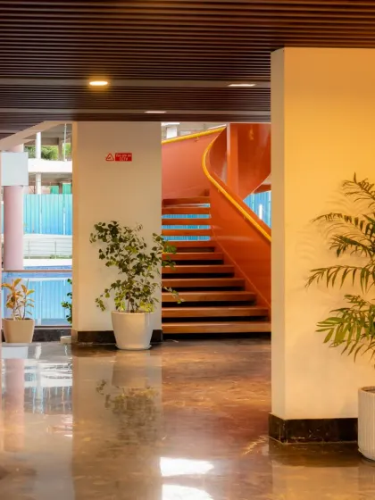 The entrance lobby of the Sona Vistaas Clubhouse, where polished marble floors reflect the warm light and greenery. This view offers a glimpse of the central staircase in the background, hinting at the dynamic spaces that unfold within.