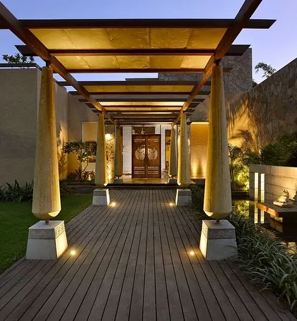 The entrance to C Farm in Chattarpur features a wooden deck walkway flanked by sculptural columns, leading to a grand door. The design uses a covered pergola to create a dramatic sense of arrival and transition from the outside world.