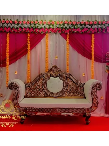 A beautiful stage setup for an engagement or reception, with a white carved sofa against a backdrop of pink drapes and floral arrangements.