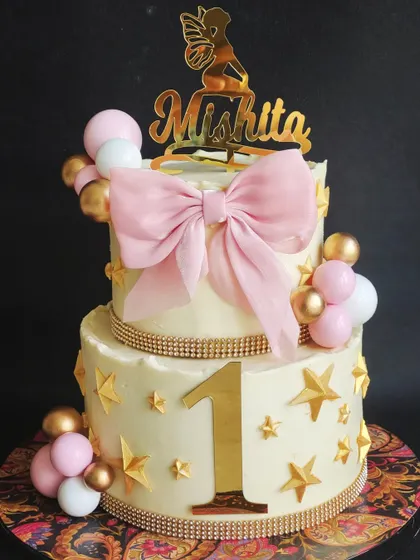 A sweet and elegant two-tiered cake for a first birthday. The design features a large edible bow, gold stars, and rhinestone ribbons for a touch of sparkle.