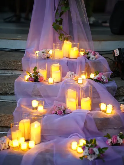 A romantic centerpiece that sets the mood. This arrangement of mixed florals in a vintage-style urn, surrounded by glowing candles on a bed of lavender fabric, creates an enchanting focal point for the table.