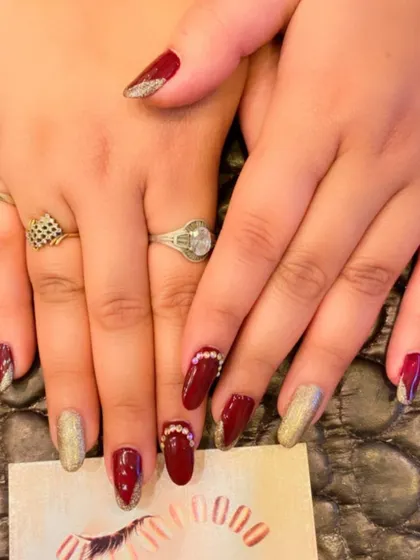 A festive and glamorous set of almond nails. The design includes deep red polish, gold glitter accent nails, and red nails with V-shaped glitter tips and rhinestone cuticle details.