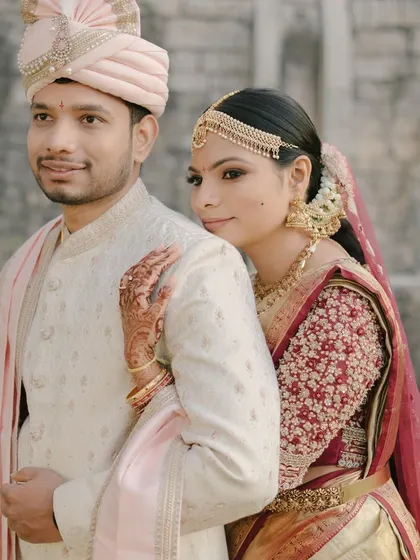 The groom's cream sherwani features subtle self-embroidery and is paired with a soft pink stole, perfectly picking up the colors in his bride's maroon and gold saree for a harmonious and elegant couple portrait.
