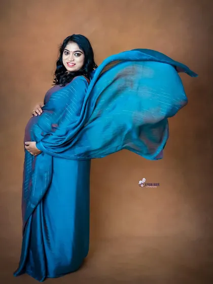 A stunning portrait of a mom-to-be in a blue saree, with the fabric tossed to create a beautiful, flowing effect against a neutral studio background.