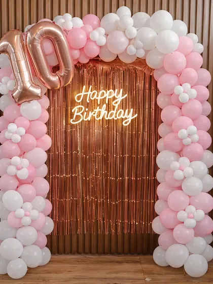A beautiful setup for a 10th birthday, featuring a pink and white balloon arch with delicate flower details. The rose gold foil curtain and "Happy Birthday" neon sign add a touch of glamour to the celebration.