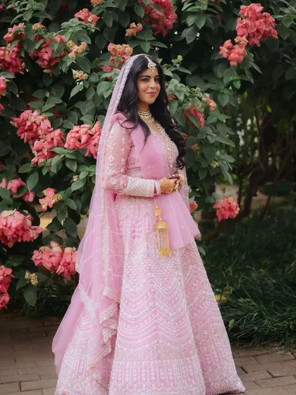 This full-length shot shows a bride looking serene and beautiful against a floral backdrop. Her pink lehenga and soft makeup look are perfect for an outdoor or garden wedding ceremony.