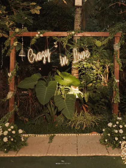 A "Crazy in Love" neon sign provides a fun and modern photo booth backdrop for an evening reception. It's set against a wall of natural greenery, blending rustic and contemporary styles.