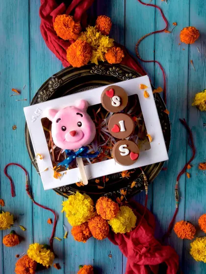 Rakhi is a special celebration of the bond between siblings. This adorable hamper for a sister includes a cute pink teddy bear cakesicle and Oreo-filled chocolates that spell out "SIS," all beautifully presented in a festive box.