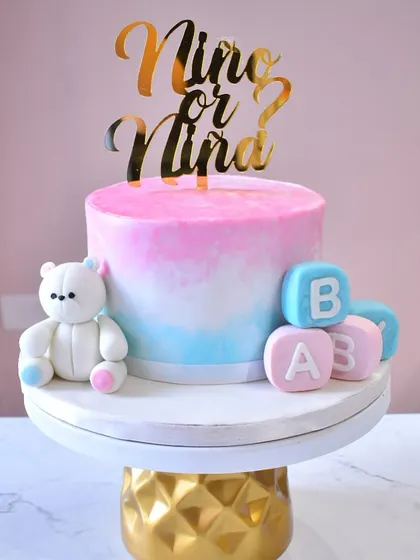 A lovely gender reveal cake with a "Niño or Niña?" theme. The cake has a pink and blue ombre effect and is decorated with a sweet teddy bear and fondant baby blocks.