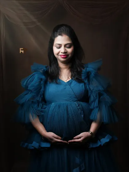A serene solo portrait. The mom-to-be cradles her bump, looking peaceful and beautiful in a tiered blue gown. The subtle lighting effects add a divine glow.