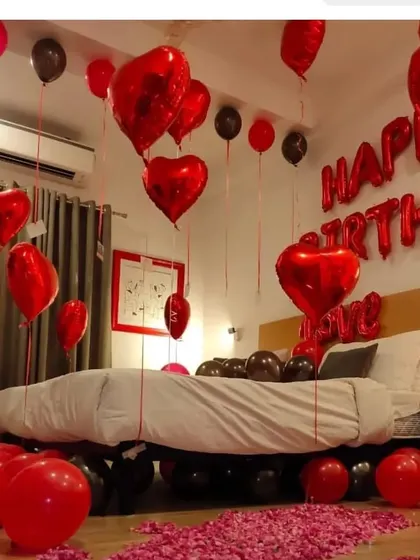 A cozy and romantic birthday room setup. The decor features red and black balloons, heart-shaped foils, and rose petals scattered on the floor and bed.