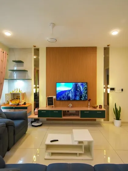 This living room features comfortable recliner sofas in a deep blue, a minimalist TV unit with green and wood accents, and a dedicated puja corner, demonstrating a blend of modern comfort and traditional values.