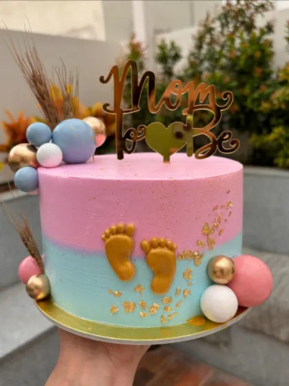 A sweet "Mom to Be" cake for a baby shower. The pink and blue colors are perfect for a gender reveal, decorated with tiny gold feet and a custom topper.