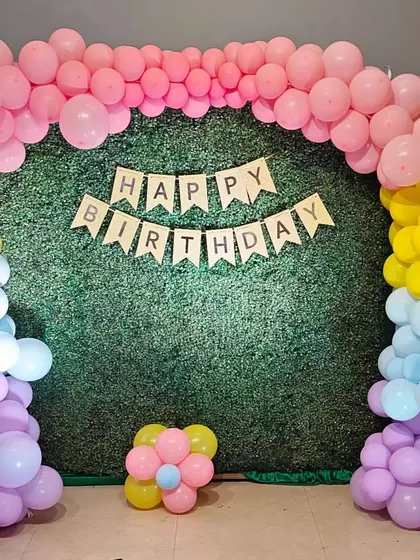 A simple birthday backdrop with a pastel balloon arch over a green hedge wall.