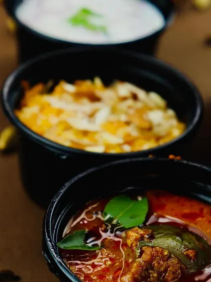 A close-up of the essential sides that complete our biryani meal. This includes our tangy brinjal gravy (Baigan ka Khatta), creamy onion raita, and a portion of rich carrot halwa for dessert.