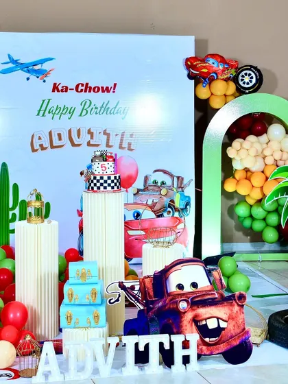 A closer look at the 'Cars' themed setup, showing the cake on a fluted plinth, a Mater cutout, and personalized 'ADVITH' block letters.