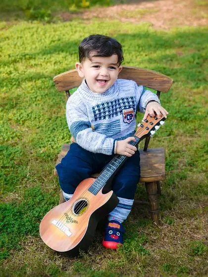 A big, happy smile from a toddler boy holding a guitar during his outdoor photoshoot. His joy is evident.