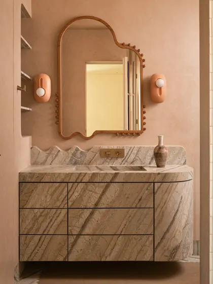 The vanity area is a beautiful composition of curves, textures, and soft colors, from the wavy marble backsplash to the organic mirror and peach-colored sconces.