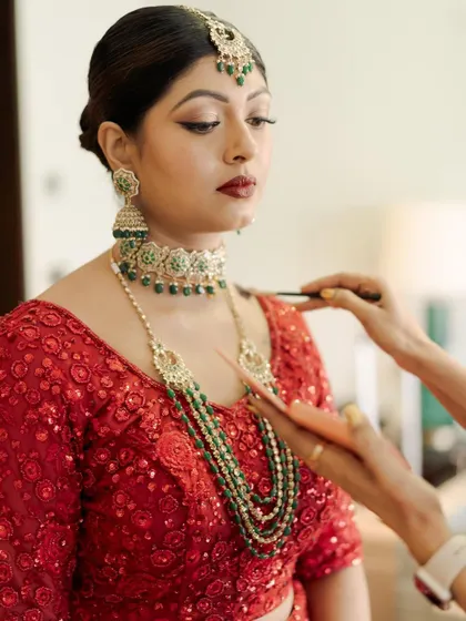 A behind-the-scenes moment, applying the finishing touches. The combination of the red outfit and green jewelry is a classic and beautiful choice.