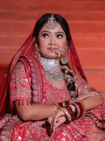 A thoughtful portrait of a bride looking upwards, seated on steps. The warm, dramatic lighting emphasizes the rich red color of her lehenga and the sparkle of her jewellery.