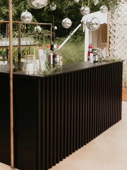 A modern and minimalist bar setup for a chic outdoor party. The black, vertically fluted bar counter is complemented by industrial-style shelving and hanging disco balls for a touch of fun.