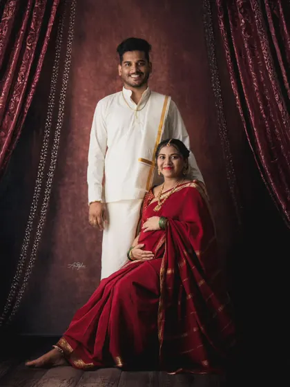 A classic portrait of a couple in traditional South Indian attire. The studio setup with rich, dark curtains creates a formal and timeless feel for this maternity photo.
