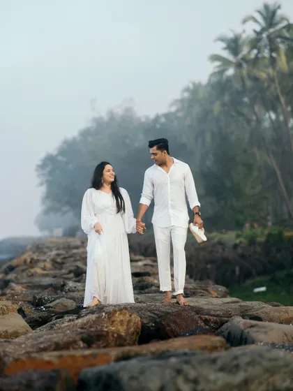 A couple enjoys a romantic walk on coastal rocks, hand in hand, with a misty, atmospheric background of the sea and trees.