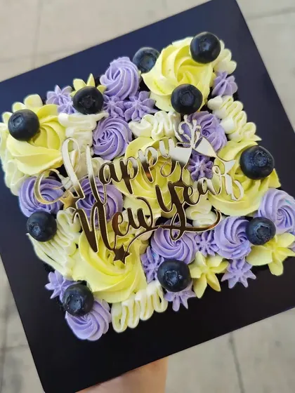A square sheet cake for the New Year, decorated with piped flowers in yellow and purple and topped with fresh blueberries.