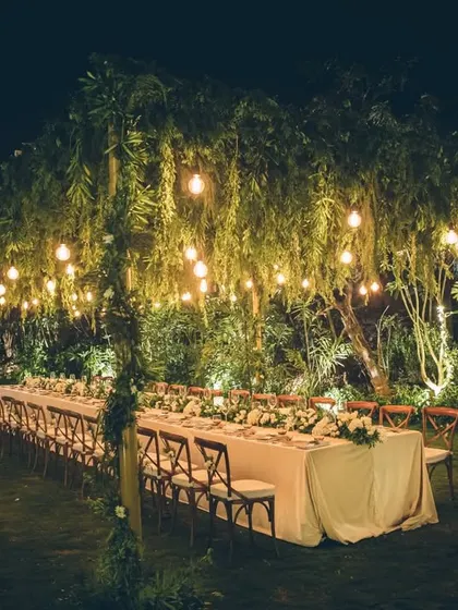 A long dining table set for an intimate dinner in a garden, under hanging lights.