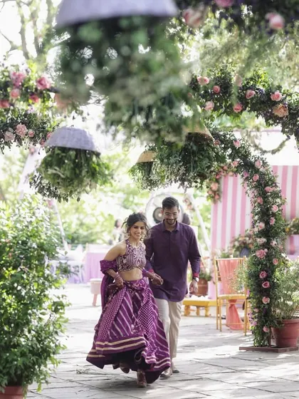 A couple walks through a romantic garden path lined with arches of pink roses.