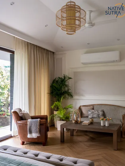 A sitting area within the master bedroom, featuring a classic sofa and a leather armchair. The space is designed for relaxation, with a large rattan pendant light and herringbone wooden floors adding warmth and texture.