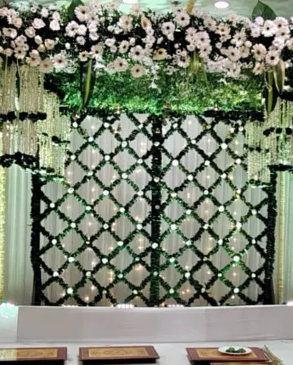 An elegant wedding ceremony stage with a backdrop made of green leaves and white flowers in a lattice pattern. The hanging floral arrangements add depth and beauty to the mandap.