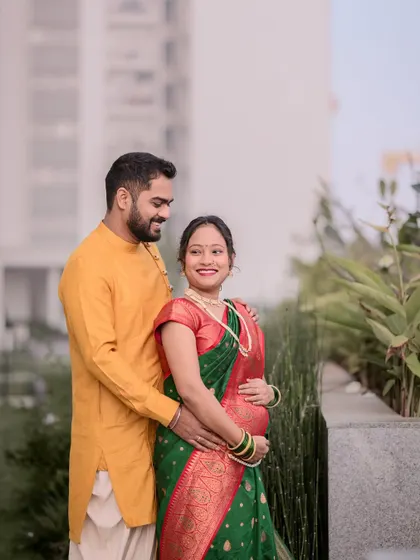 A lovely outdoor portrait of an expecting couple. The husband-to-be has his arm around his wife, who is radiant in a traditional green and red saree during their maternity session.
