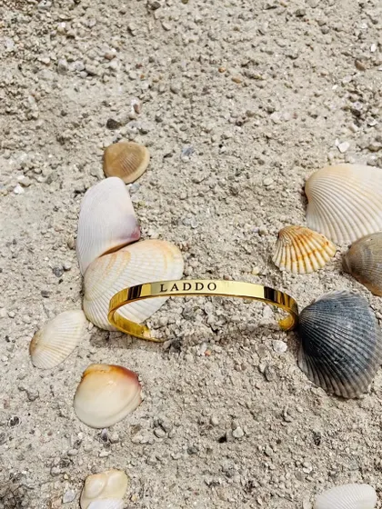 A custom gold-plated Kada with the nickname 'Laddo' engraved, resting on the sand among seashells. A perfect, sunny shot of a personal treasure.