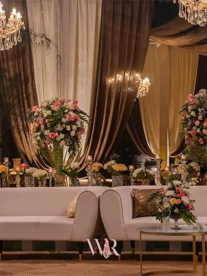 A luxurious lounge area with plush white sofas, modern coffee tables, and large floral arrangements. The backdrop of rich brown and cream drapes, along with the chandeliers, creates a sophisticated and comfortable setting.
