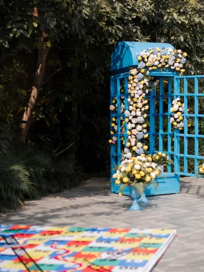A quirky photo-op featuring a vintage British telephone booth, painted bright blue and overflowing with flowers, set next to a giant snakes and ladders game board.