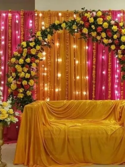 A bright and cheerful Haldi or Mehndi decoration for home. The combination of pink and orange drapes with a floral arch and fairy lights creates a warm and inviting backdrop for your ceremony.