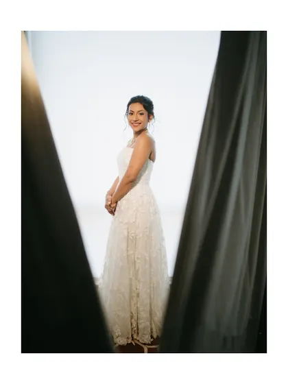A simple and beautiful portrait of the bride in her wedding gown, framed by curtains. The soft light gives her an angelic glow.