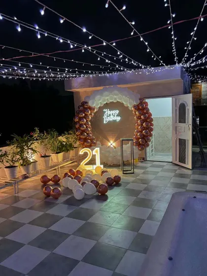 A 21st birthday celebration on a rooftop terrace. The setup includes a circular backdrop with a neon sign, marquee numbers, and fairy lights strung overhead for a magical ambiance.
