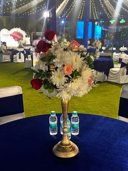 A close-up of a table centerpiece at a blue and white themed event. The arrangement of red roses and white chrysanthemums in a brass vase adds a touch of elegance to the guest tables.