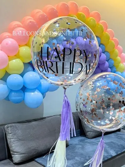 A pastel rainbow balloon arch over the sofa creates a dreamy backdrop for a birthday celebration, complemented by personalized confetti balloons.