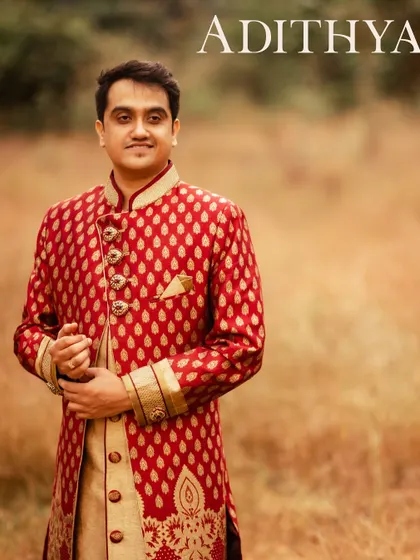 A handsome portrait of the groom in a striking red and gold sherwani, set against a soft, natural background. This outdoor shot uses natural light to highlight the intricate details of his traditional attire.